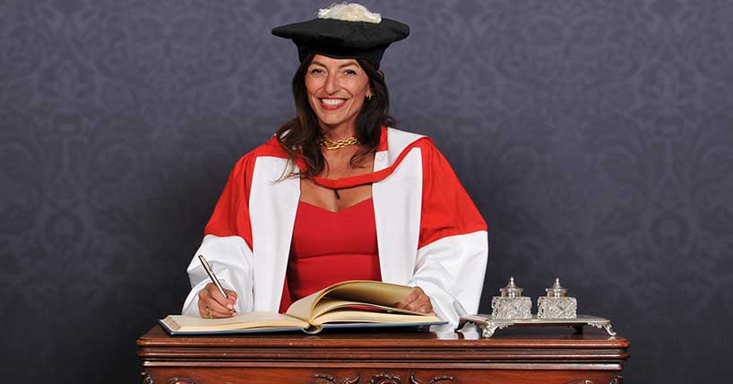 Davina McCall in academic robes sitting at a desk