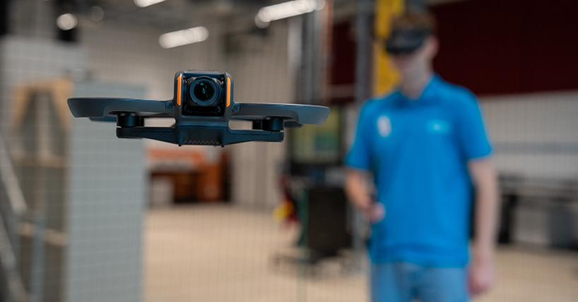 A student in a blue t shirt with a headset on guiding a drone flying in the foreground A student in a blue t shirt with a headset on guiding a drone flying in the foreground