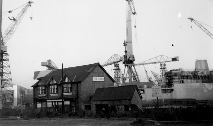 B&W photo of the Dock pub in Wallsend, Tyne and Wear, taken in the late 1980s. A ship and several of the iconic Swan Hunter shipyard cranes tower above the pub.
Andrew Green