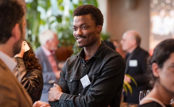 Young dark-skinned man in a dark shirt, smiling with arms crossed, engaged in conversation at an alumni networking event. Other attendees, both men and women, are visible in the background. The setting includes plants and a well-lit modern interior.

