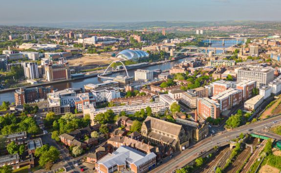 A wide aerial view of Newcastle upon Tyne, featuring the River Tyne, the Glasshouse International, the Millennium Bridge, and surrounding buildings and infrastructure on a clear day.