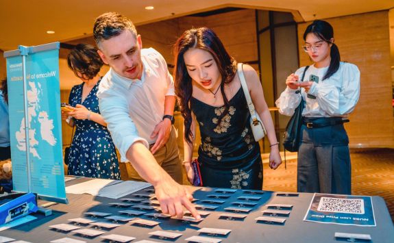 Light-skinned man assisting a light-skinned woman at an information booth, pointing to name tags on a table at an alumni event in China. 