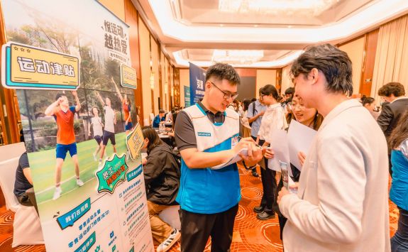 Two light-skinned individuals discussing at a booth with a poster display, featuring Chinese text about health and fitness.