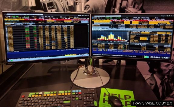 Dual monitors displaying financial data on a Bloomberg terminal, including market statistics, price trends, and volume analysis, with a keyboard and control panel in the foreground.