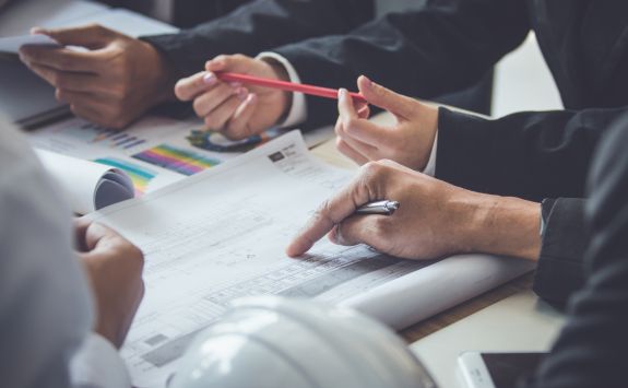 Close-up of hands of business professionals reviewing architectural blueprints and charts, with one person pointing at the plans and another holding a red pencil.