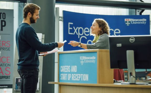 A male student with light skin and curly hair is receiving papers from a woman with light skin and curly hair at the Careers and Start-up Reception desk at Newcastle University. The background features banners with university branding.