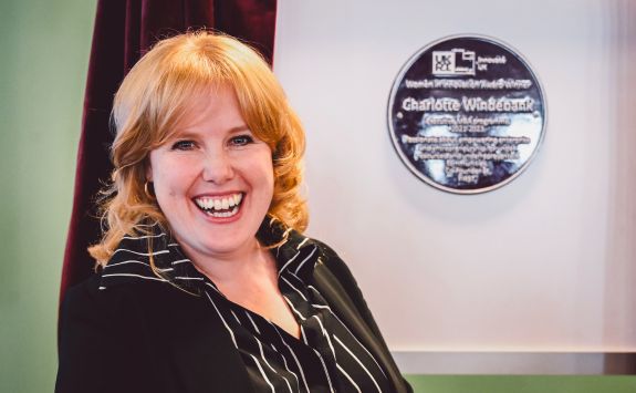 Charlotte Windebank smiling at her Women in Innovation Award ceremony, standing in front of her award plaque recognizing her work in empowering underrepresented communities.