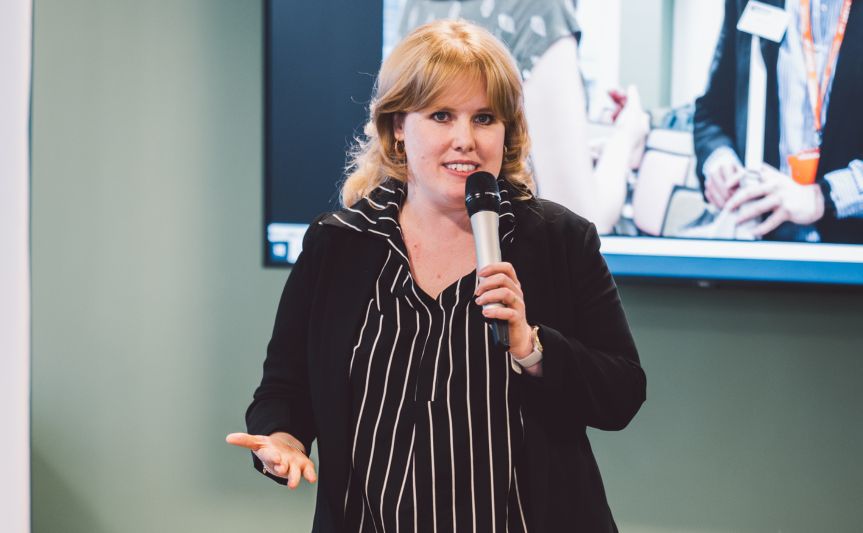 Charlotte Windebank speaking at the Women in Innovation Award ceremony, holding a microphone and addressing the audience with a presentation screen behind her.