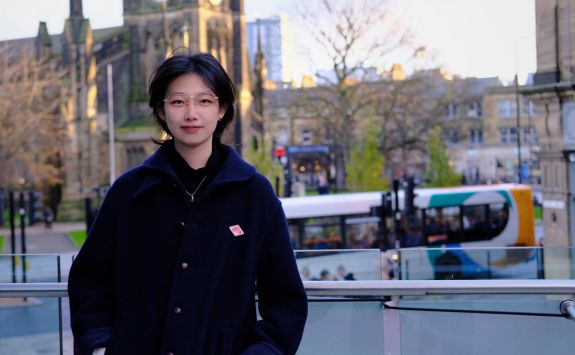 Congjing Song, a light-skinned woman with short dark hair and glasses, standing outdoors in Newcastle with historic buildings and a passing bus in the background.