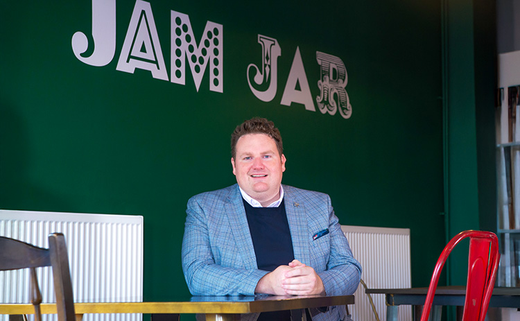 Dan Ellis, a light-skinned man with short curly hair, sits at a table in Jam Jar Cinema, smiling at the camera. The green wall behind him features the venue