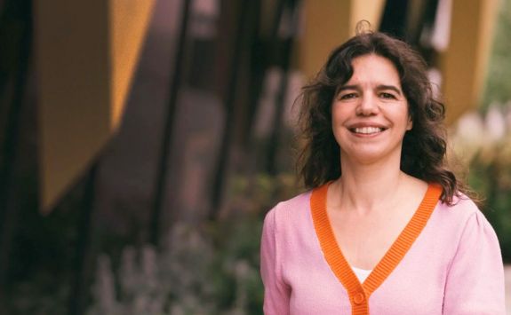 Dr Ana Lopes smiling while standing outdoors in front of a building. She is wearing a pink cardigan with an orange trim.