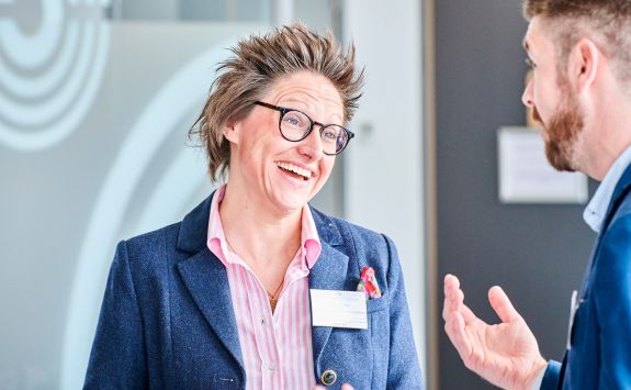 Dr Emily Yarrow wearing a blue blazer and glasses, smiling and talking to a colleague in a modern office setting.