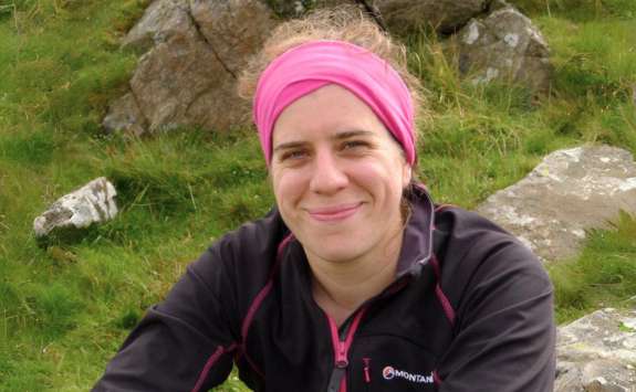 Dr Emma Hill wearing a pink headband and a black jacket with pink details, smiling in an outdoor setting with rocks and greenery in the background.