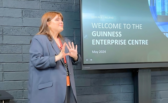 A woman in a blazer is presenting in front of a screen displaying the text "Welcome to the Guinness Enterprise Centre, May 2024" in Dublin, Ireland.