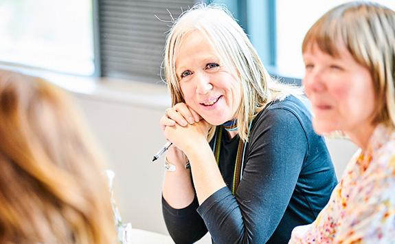 A light-skinned woman with long blonde hair smiling and holding a pen, engaged in a classroom discussion during an executive education session. Two other women, also light-skinned, sit beside her, partially visible. The background shows a bright, well-lit room.