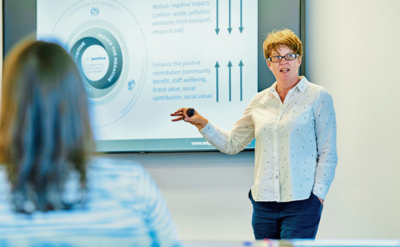 A woman presenting during an executive education session. She is standing in front of a screen displaying a circular diagram and pointing towards it while engaging with participants. A blurred participant in the foreground listens attentively.