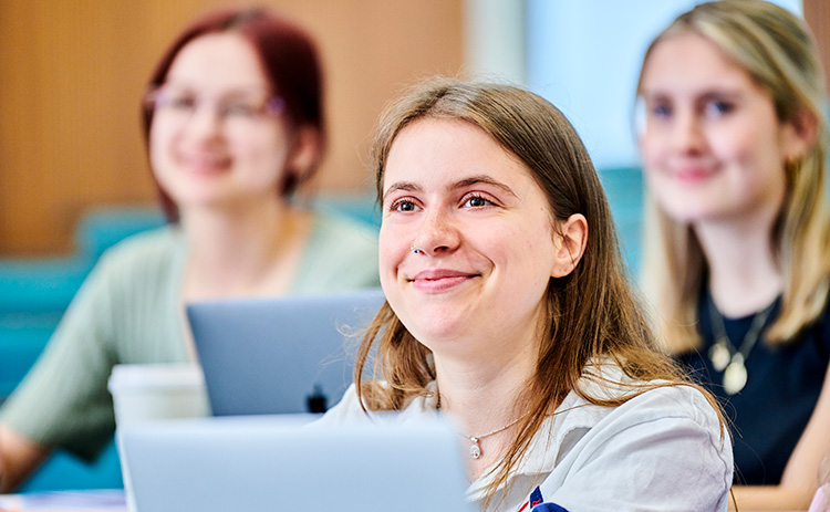 Three light-skinned female students sitting in a lecture, smiling and focused, with laptops and notebooks in front of them.	