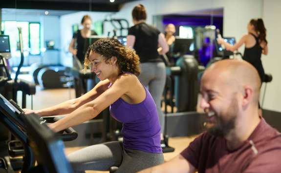 A woman and a man exercising on stationary bikes in a gym setting, the woman is smiling and appears to be enjoying her workout.