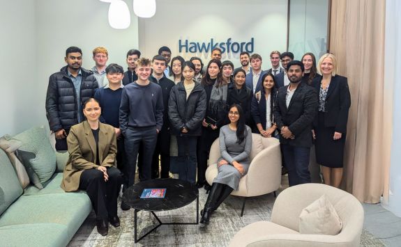 A group of students visiting Hawksford as part of the student trek. They are standing and sitting in a corporate lounge area, with the Hawksford logo on the wall behind them.