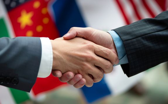 Close-up of two people in business suits shaking hands, with international flags including those of China, Italy, Russia, and the United States in the background, symbolising global business partnerships.