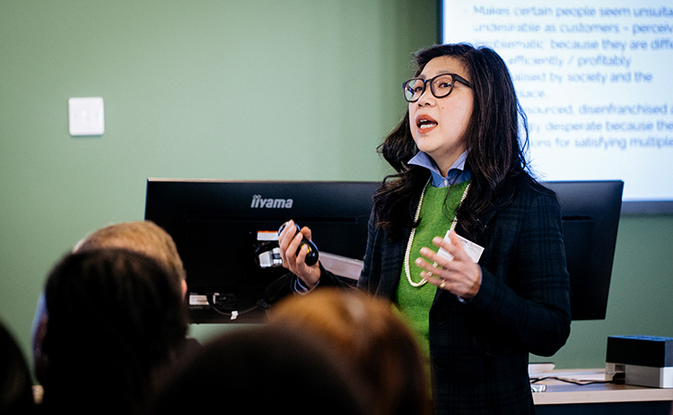 Dr Josephine Go Jefferies, wearing glasses, a green jumper, and a blazer, delivering a presentation to an audience in a classroom setting. A computer monitor and presentation screen are visible in the background.