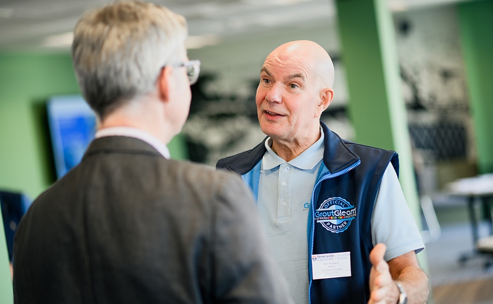 Karl Hodgekiss, wearing a light blue polo shirt and a GroutGleam branded vest, speaking with another attendee at the Newcastle University Business School (NUBS) Business Open Day. The setting features a modern green and black interior with a professional atmosphere.