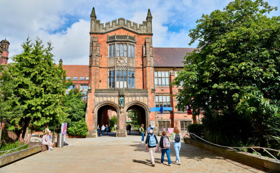 Kings Walk in Newcastle upon Tyne: A historic red brick building with a prominent archway and intricate stonework. Several people walk towards the archway, with trees and greenery lining the path.