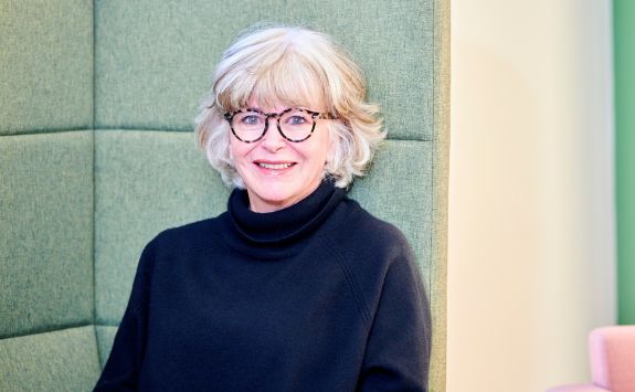 Lisa Opie, wearing a black turtleneck sweater and glasses, smiles while sitting against a green upholstered wall. 