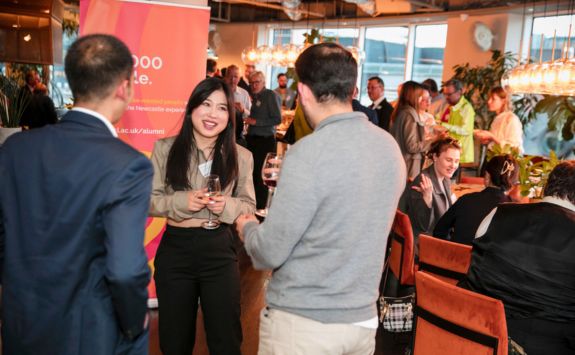 Attendees at a London alumni event are networking in a lively and elegant setting. A woman holding a drink smiles and converses with two men. The room is filled with people engaged in discussions, with a warm ambiance created by stylish lighting and decor. A banner is visible in the background.