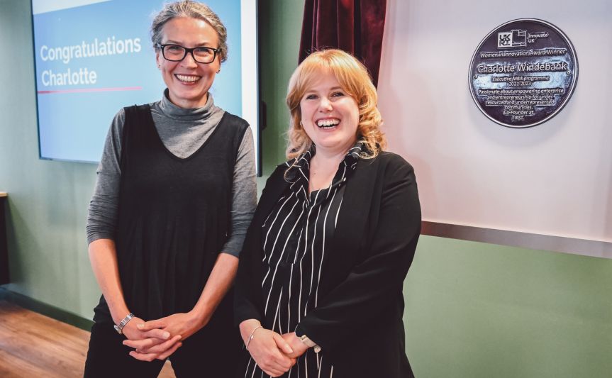  Lucy Hatt and Charlotte Windebank smiling at the Women in Innovation Award ceremony, with the plaque displayed on the wall in the background.