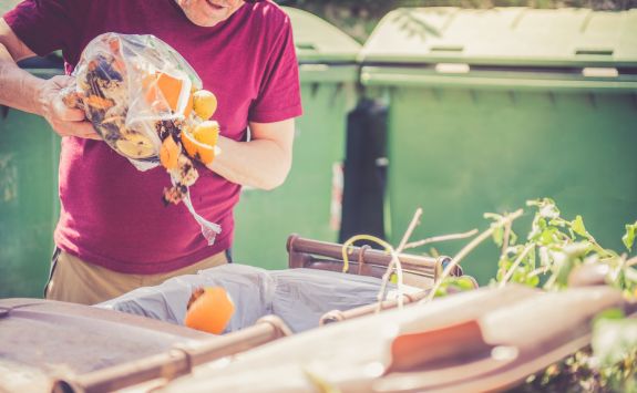 Light-skinned man wearing a red T-shirt emptying food waste into a compost bin, with green dumpsters in the background.