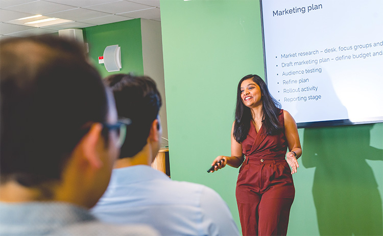 A dark-skinned female student in a burgundy jumpsuit presents a marketing plan to a group, with a projected slide behind her.