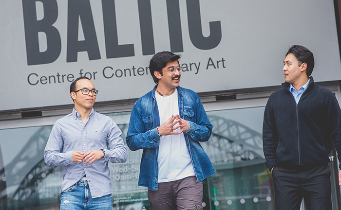 Three MBA students standing outside the BALTIC Centre for Contemporary Art in Newcastle. One student in glasses and a light blue shirt, another in a denim jacket with a moustache, and a third in a black jacket are engaged in conversation. The Tyne Bridge is visible in the background.