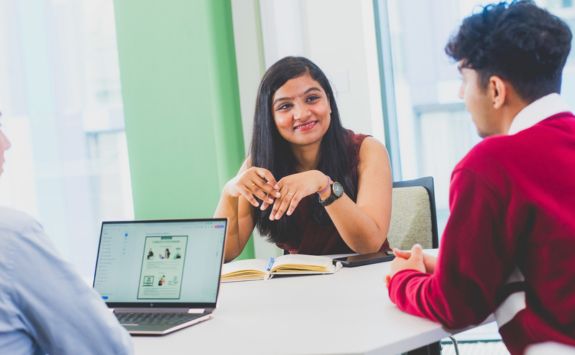 A diverse group of MBA students having a discussion around a table. A woman with a medium-brown skin tone is smiling and leading the conversation, while the others listen attentively. A laptop is open on the table.