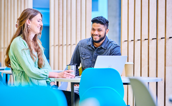 Two MBA students sitting at a table, engaged in conversation. The woman on the left, with light skin and long brown hair, smiles while holding a cup. The man on the right, with medium-dark skin, short black hair, and a beard, is working on a laptop. They are seated in a bright, modern setting with blue chairs and wooden panel walls in the background.