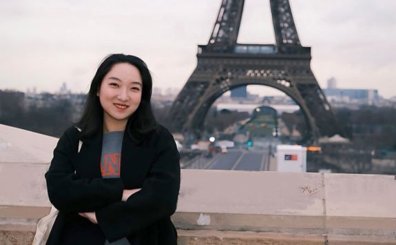 Mengya Wu, a light-skinned woman with long dark hair, smiling while standing in front of the Eiffel Tower in Paris. She is wearing a black coat and a grey sweater.