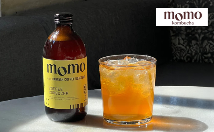 A bottle of Momo Coffee Kombucha with a yellow label sits on a marble table next to a glass filled with iced kombucha. The background features a dark sofa with patterned cushions. The 