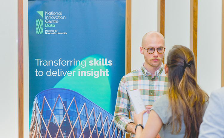 A bald male professional wearing glasses and a checkered shirt talking to a woman with long brown hair at a networking event in front of a National Innovation Centre for Data banner.