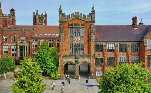 A view of the iconic arches at Newcastle University, with people walking through the entrance. The historic red-brick building is surrounded by green trees, and the sky is clear.