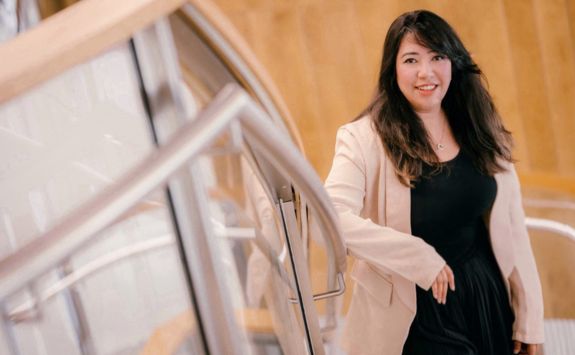 Nida Shamim, a medium-skinned woman with long dark hair, smiling while walking up a staircase. She is wearing a beige blazer over a black dress.