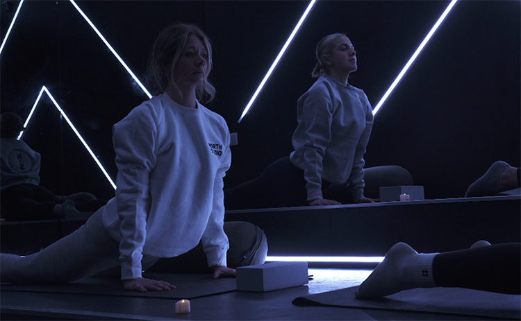 Two women in a dimly lit fitness studio practicing yoga. They are wearing sweatshirts and leggings, stretching in a cobra pose on yoga mats. The room features neon LED light strips on dark walls, a mirrored surface reflecting the participants, and yoga props like blocks and candles.