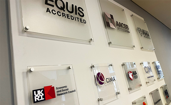 Wall display featuring accreditation plaques at Newcastle University Business School, including prominent labels such as EQUIS Accredited, AACSB Accredited, and AMBA Accredited, alongside other professional recognition plaques. The clear acrylic plaques are mounted on a white wall.