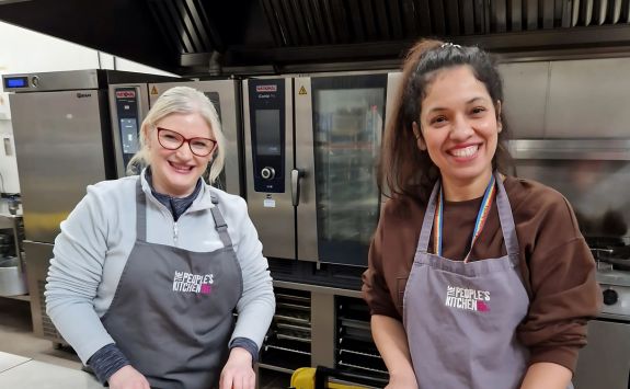 Two women smiling in a commercial kitchen setting, both wearing aprons with "The People’s Kitchen" logo. The woman on the left (Sue) has light skin, blonde hair, and glasses. The woman on the right (Sonia) has medium skin and dark hair.	