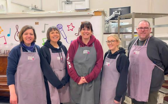 Five volunteers standing together, all wearing aprons with "The People’s Kitchen" logo. The group includes three women with light skin, one with medium skin, and one man with light skin, all smiling in a friendly indoor setting.