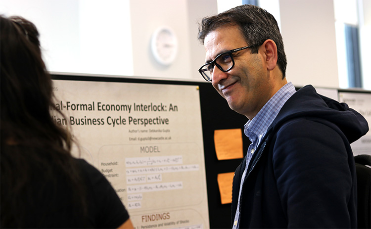 A male academic with light skin wearing glasses, smiling and engaging with a female student in front of a research poster. The poster discusses an economic model.