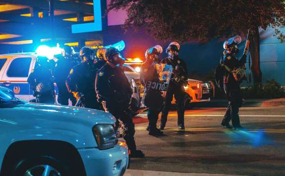 Police in riot gear walking past vehicles at night with emergency services lights in the background