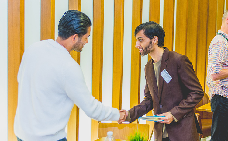 Postgraduate student with light brown skin and short dark hair, wearing a brown blazer, shaking hands with another person with light skin and dark slicked-back hair, wearing a white jumper.