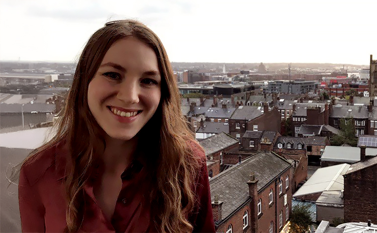 Rebecca McEwen-Orr wearing a burgundy blouse smiles while standing outdoors. The background shows a cityscape with buildings and rooftops in Newcastle, under an overcast sky.