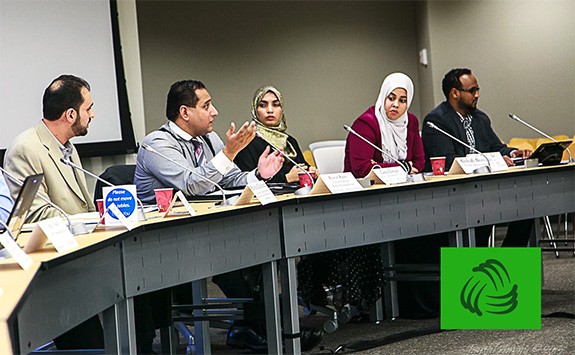 A diverse group of professionals engaged in a discussion at a conference table. The panel includes men and women, some wearing headscarves, seated with microphones, nameplates, and documents. 