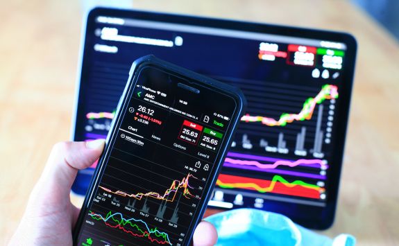 A hand holding a smartphone displaying stock market data with a tablet in the background showing financial charts.